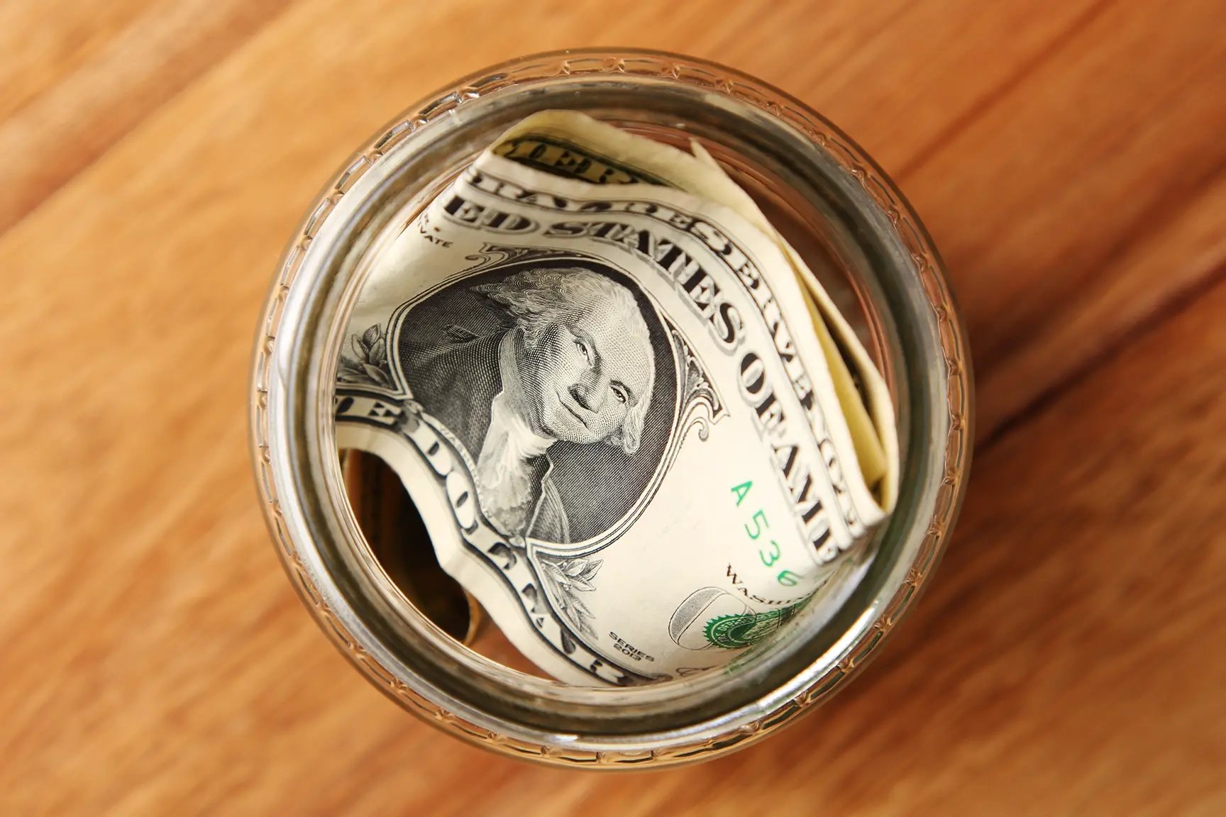 US dollar bills in a glass tip jar on a wooden counter.