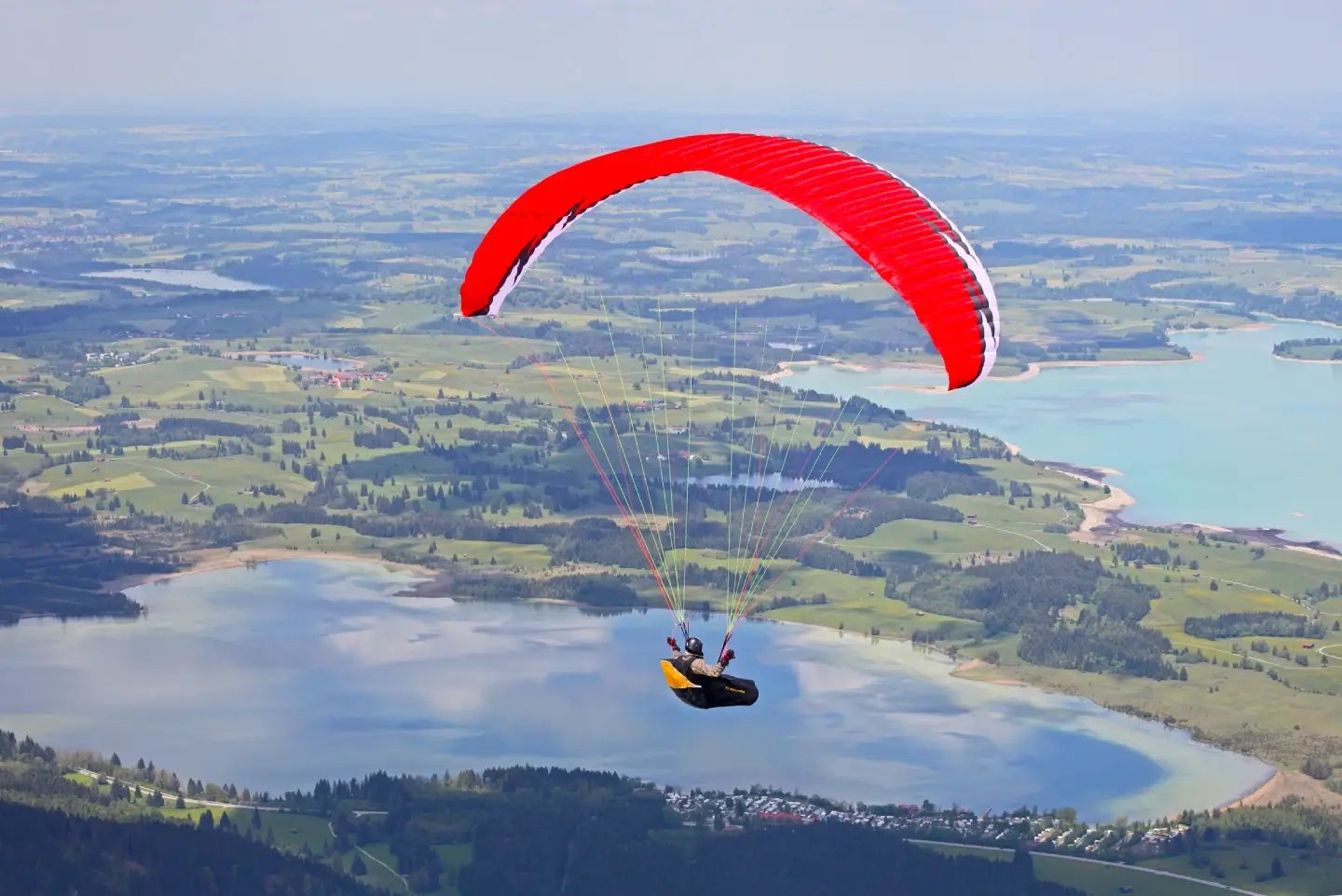 A paraglider in a red canopy soaring above a lake and surrounding hills.