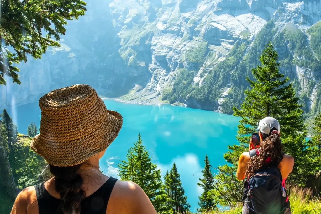 Two hikers in straw sun hats looking out over Oeschinen Lake's turquoise waters, surrounded by pine trees and the peaks of the Jungfrau-Aletsch region in the Swiss Alps, Switzerland.