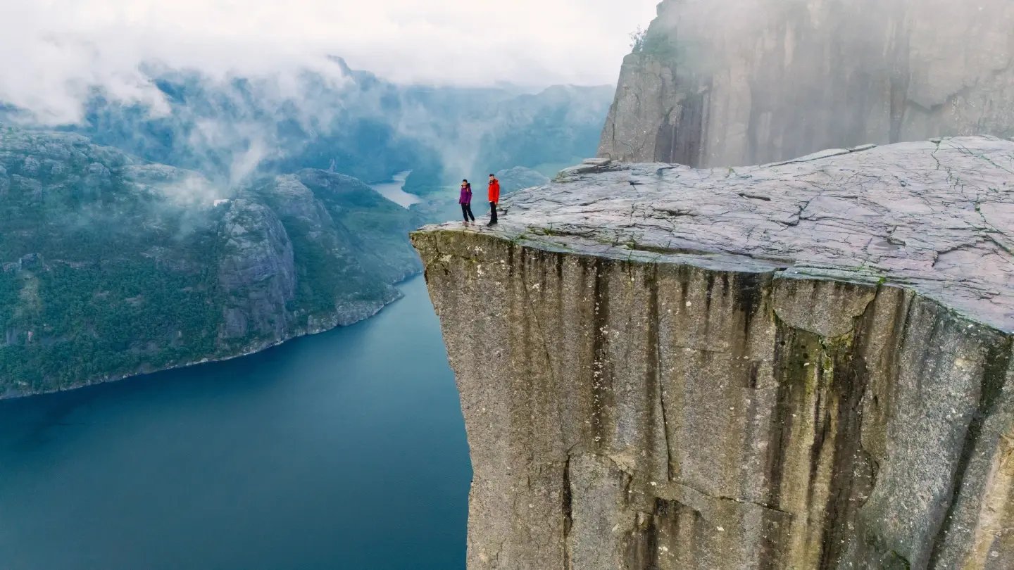 Two hikers standing on the edge of Preikestolen (Pulpit Rock) cliff overlooking Lysefjord, Norway.