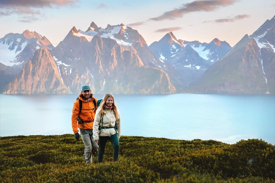 A couple in warm hiking layers standing in golden tundra grass, with a fjord and dramatic snow-capped mountain peaks behind them, likely on Senja Island in Northern Norway.