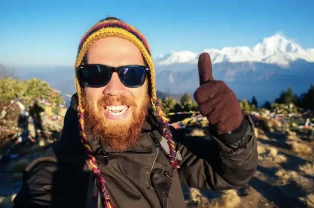 A trekker in cold-weather gear giving a thumbs up with snow-capped Himalayan peaks behind him, Nepal.