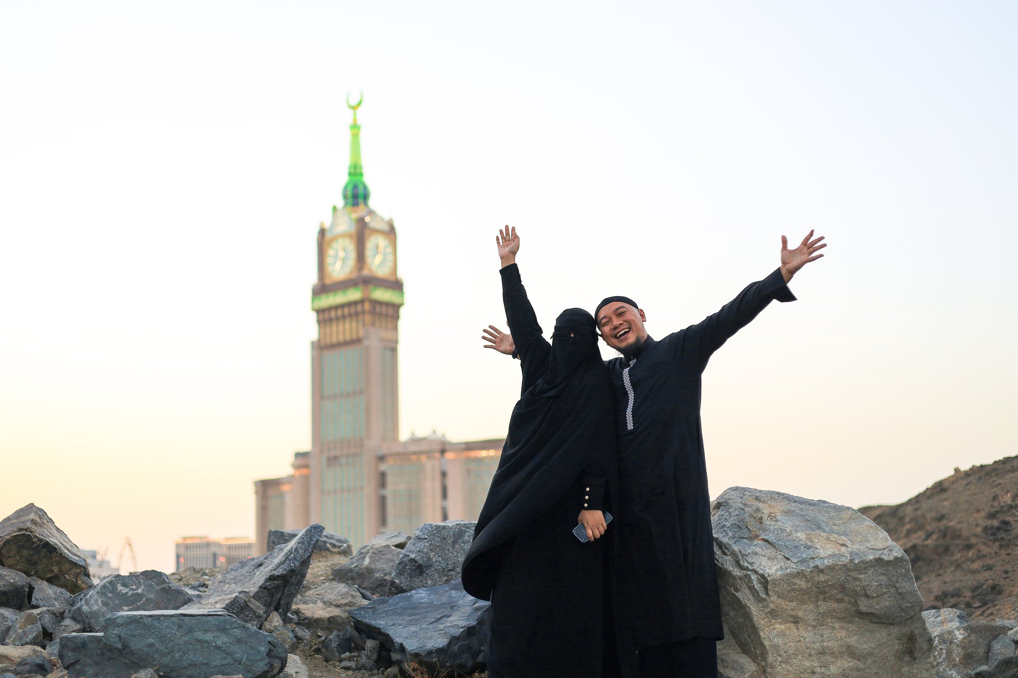 Two travellers waving in front of the Makkah Royal Clock Tower