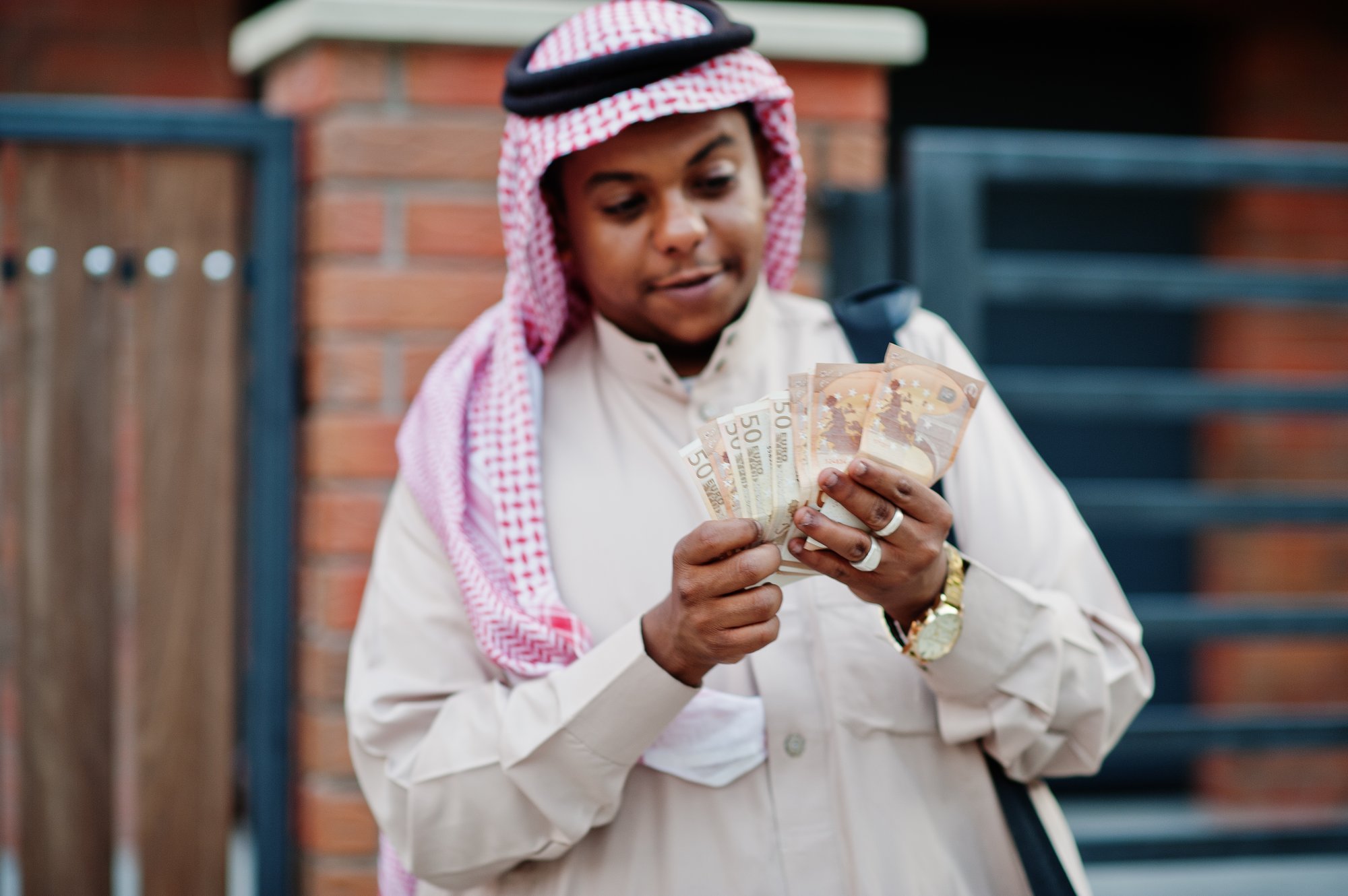 Man in a red-checked shemagh counting Saudi riyal banknotes